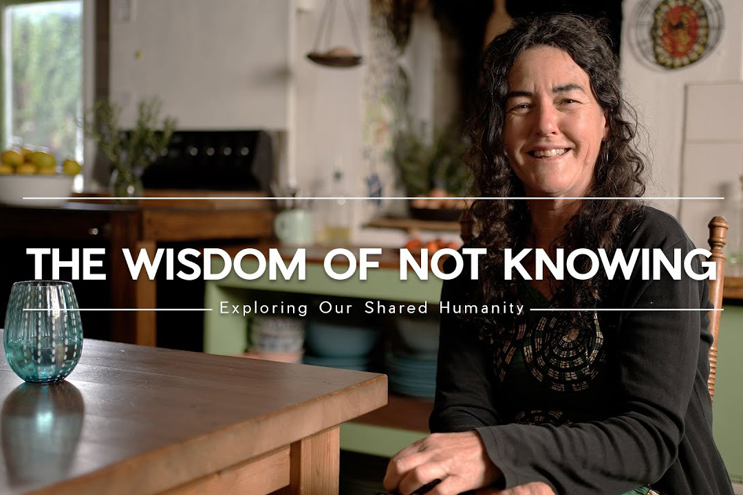 Woman with curly hair wearing a black shirt and smiling sitting at kitchen table with text that reads, "The Wisdom of Not Knowing"